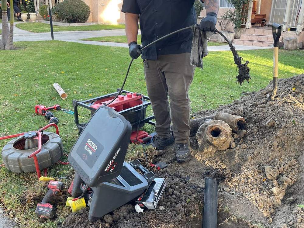 FastFlow technician feeding a sewer camera into a dug trench, with the rooter cable and a pulled root mass